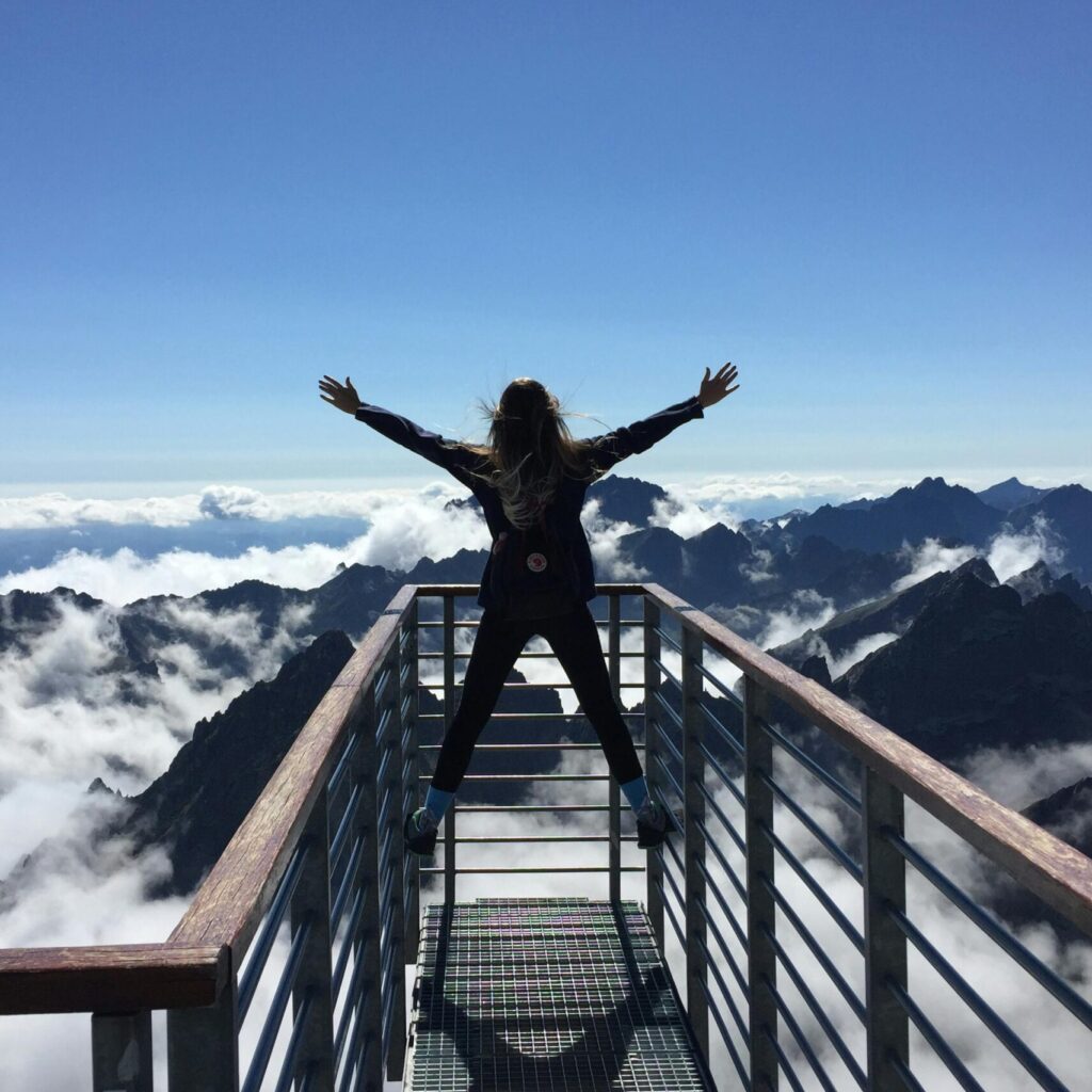 A woman stands on a viewing platform in Vysoké Tatry, Slovakia, surrounded by clouds and mountains, embracing freedom.