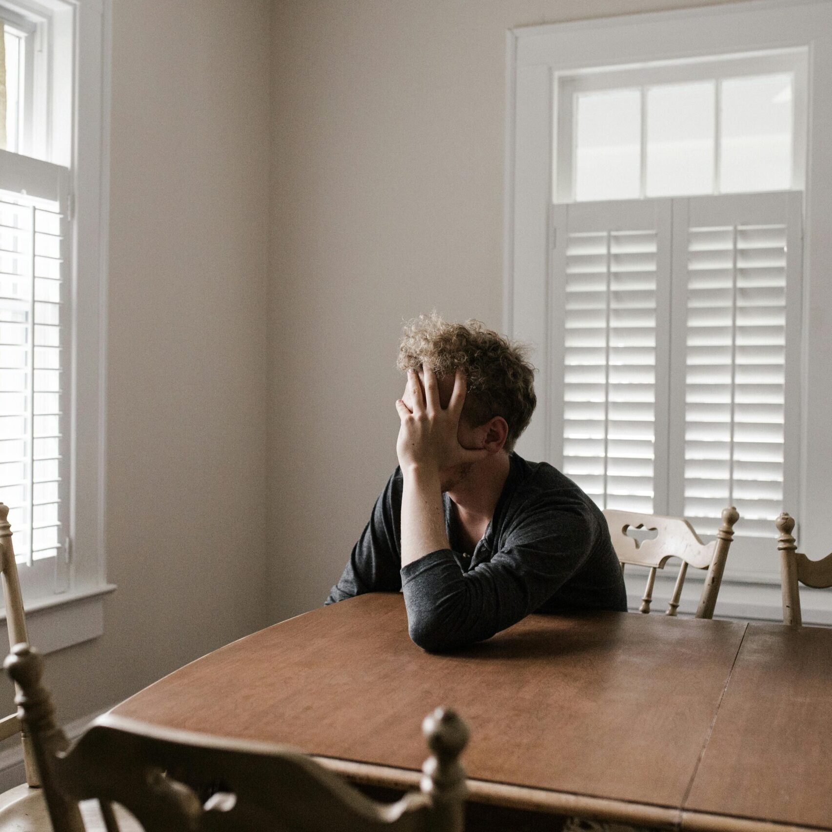 A man sits alone at a table in a bright room, displaying deep contemplation.