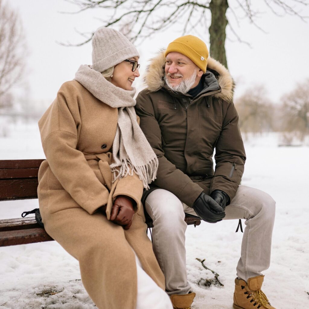 Happy elderly couple sitting on a snowy bench, wrapped in winter coats and sharing a joyful moment.