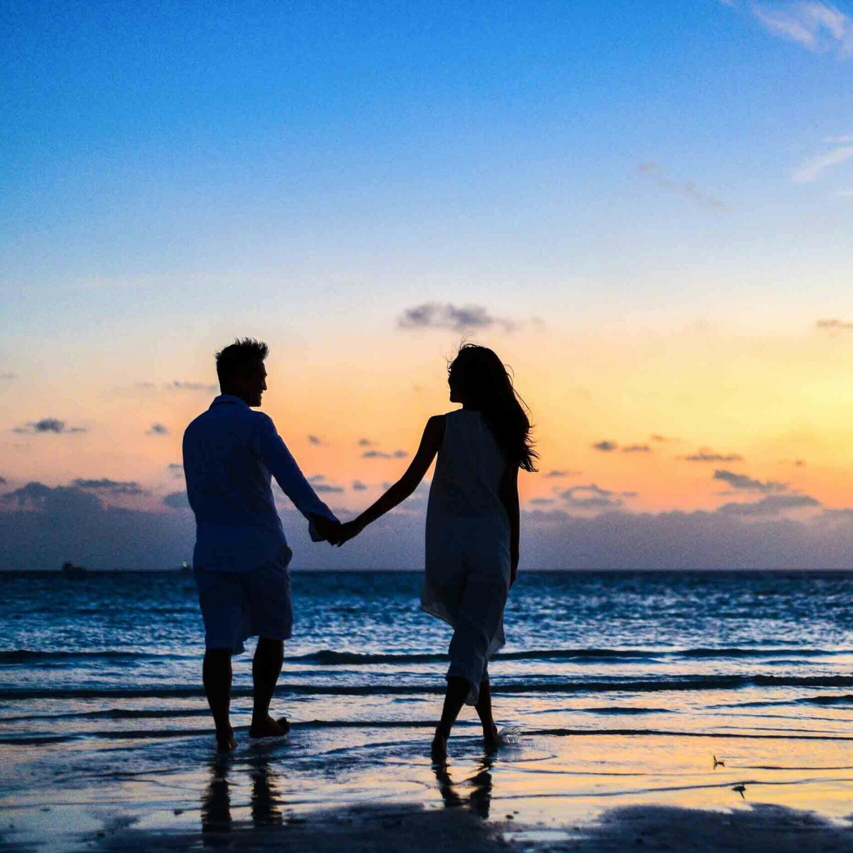 A romantic couple walks hand in hand on a tropical beach at sunset, enjoying a serene moment together.